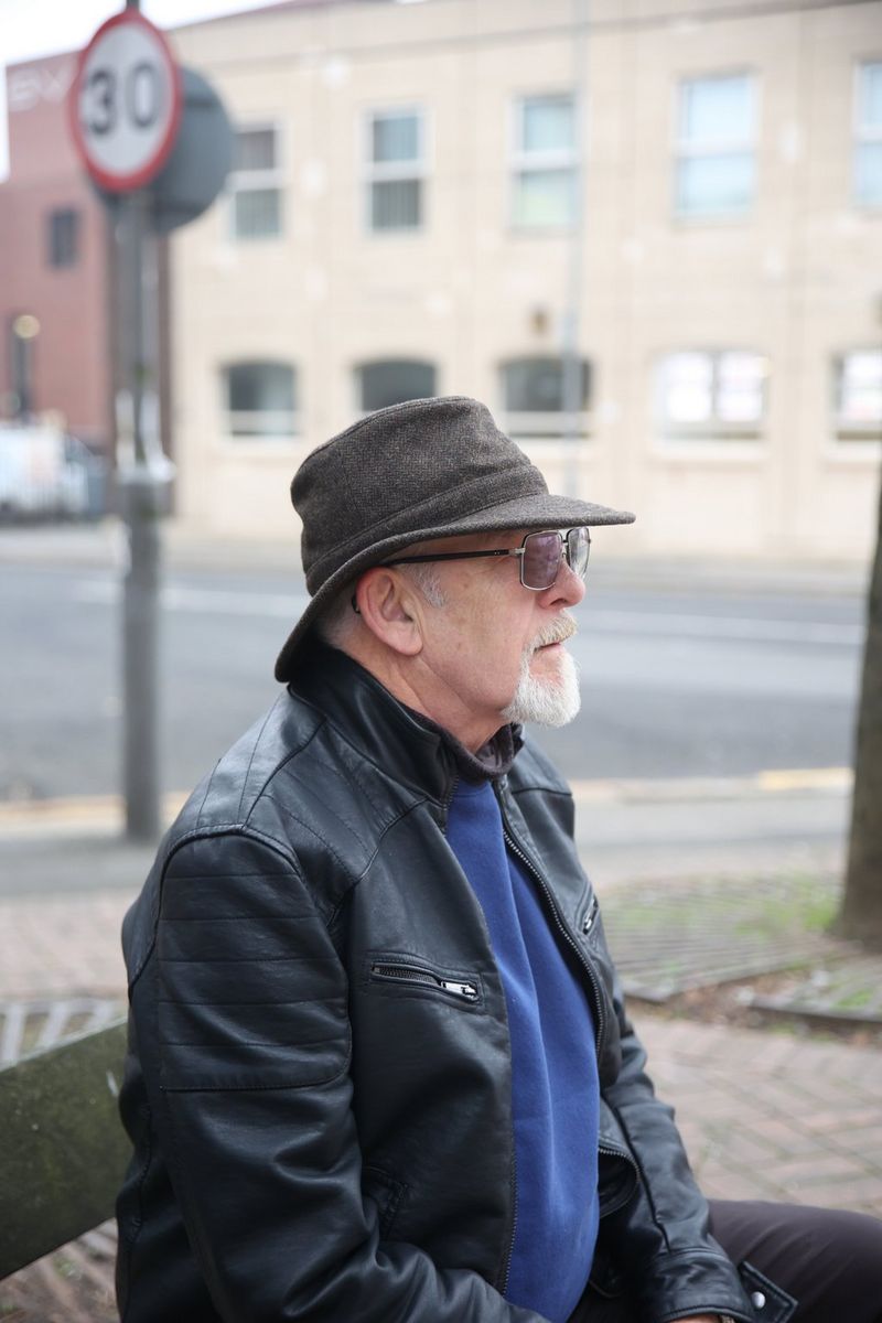 A man with a neat white beard, wearing a leather coat, tinted glasses and a tweedy hat, sits on a bench side on to the camera.
