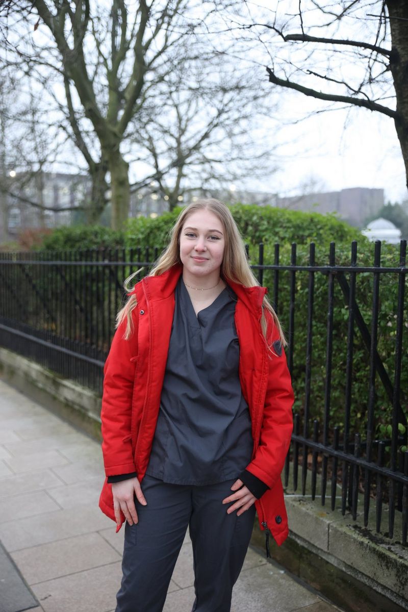 A woman with long blonde hair, wearing a bright red coat over black scrubs, stands alongside a black iron fence, looking at the camera.
