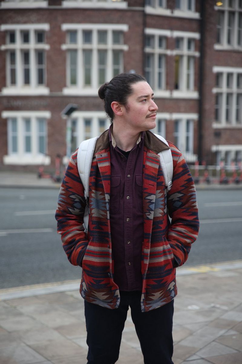 A man in a striking patterned jacket and dark hair in a bun stands with his hands in his coat pockets and his head turned to the side.