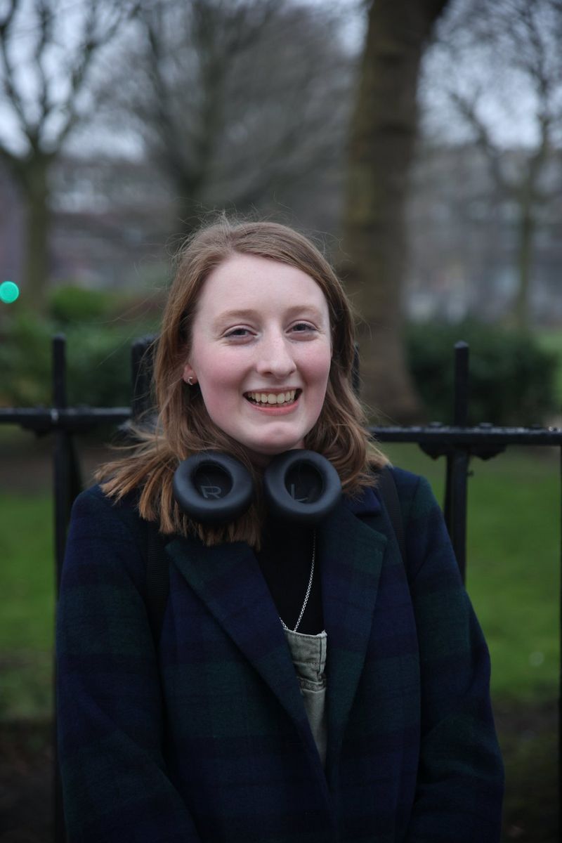 A young woman with long brown hair wearing a heavy overcoat and with large headphones around her neck smiles cautiously at the camera.