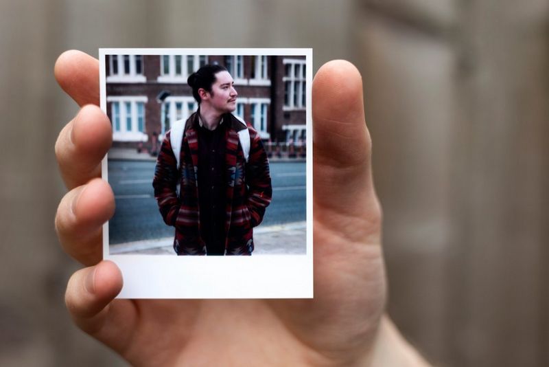 A hand holds up a small printed portrait of a young man wearing a striking patterned jacket with his head turned to the side.