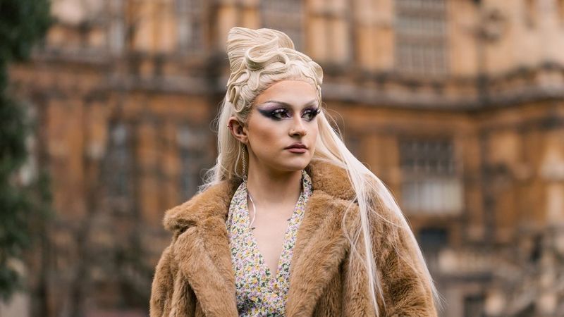 A portrait taken by Louis Painter of a drag queen, with long blonde hair and wearing a floral dress and brown fur coat, standing in front of an ornate building.