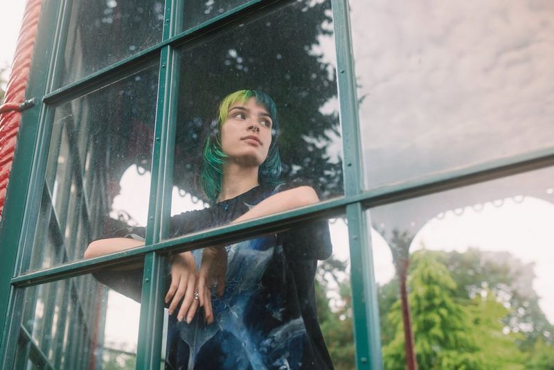 A person stands looking through a window, their brightly coloured hair complementing the greens of the window frame.