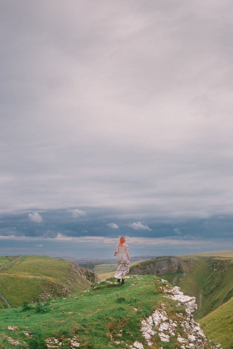 A figure in a long dress stands on the edge of a rocky hilltop, facing out towards the fields and water in the distance.