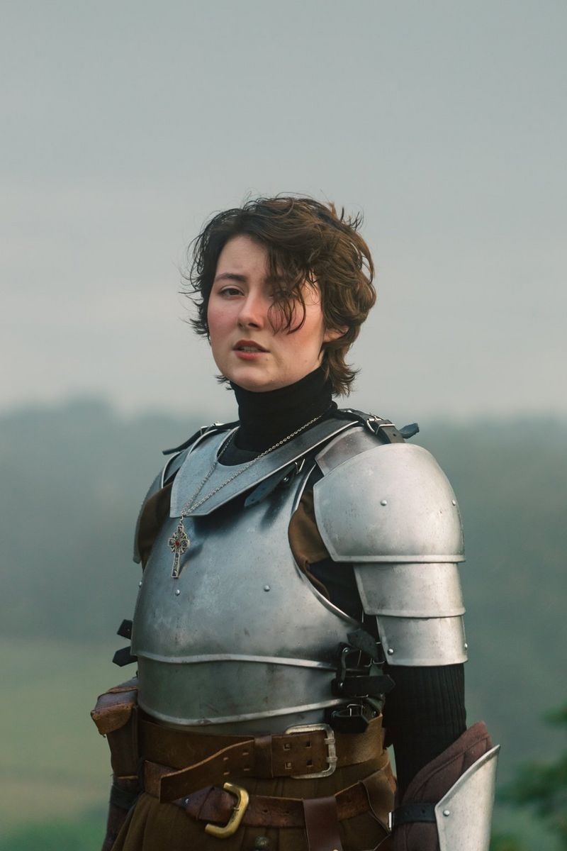 A figure stands dressed as a knight in metal armour, looking towards the camera, the background out of focus behind them.