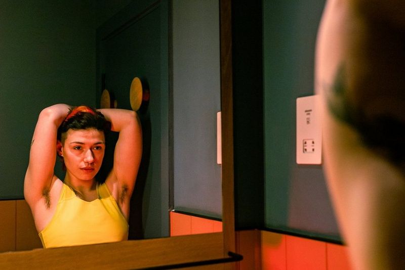 Trans musician Jay Page looks into a mirror, their hands behind their head, in a photograph taken by Louis Painter.