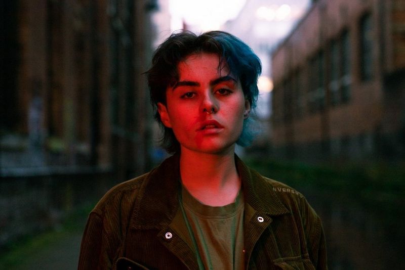 A person with short dyed hair and a nose ring looks towards the camera.