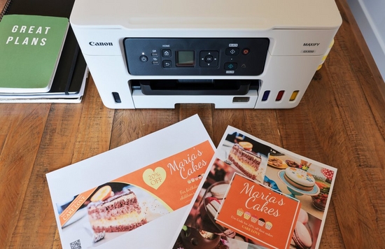 Printed Canon PosterArtist designs on a desk in front of a Canon MAXIFY printer. 