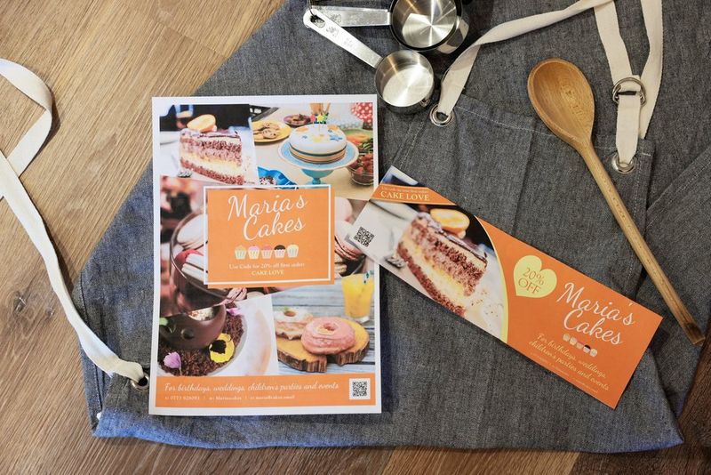 A printed poster and banner promoting a cake-making business arranged on a grey apron alongside measuring cups and a wooden spoon. 