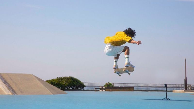 A skateboarder mid-jump against a bright blue sky. A Canon PowerShot PX is mounted on a tripod to the right of him.