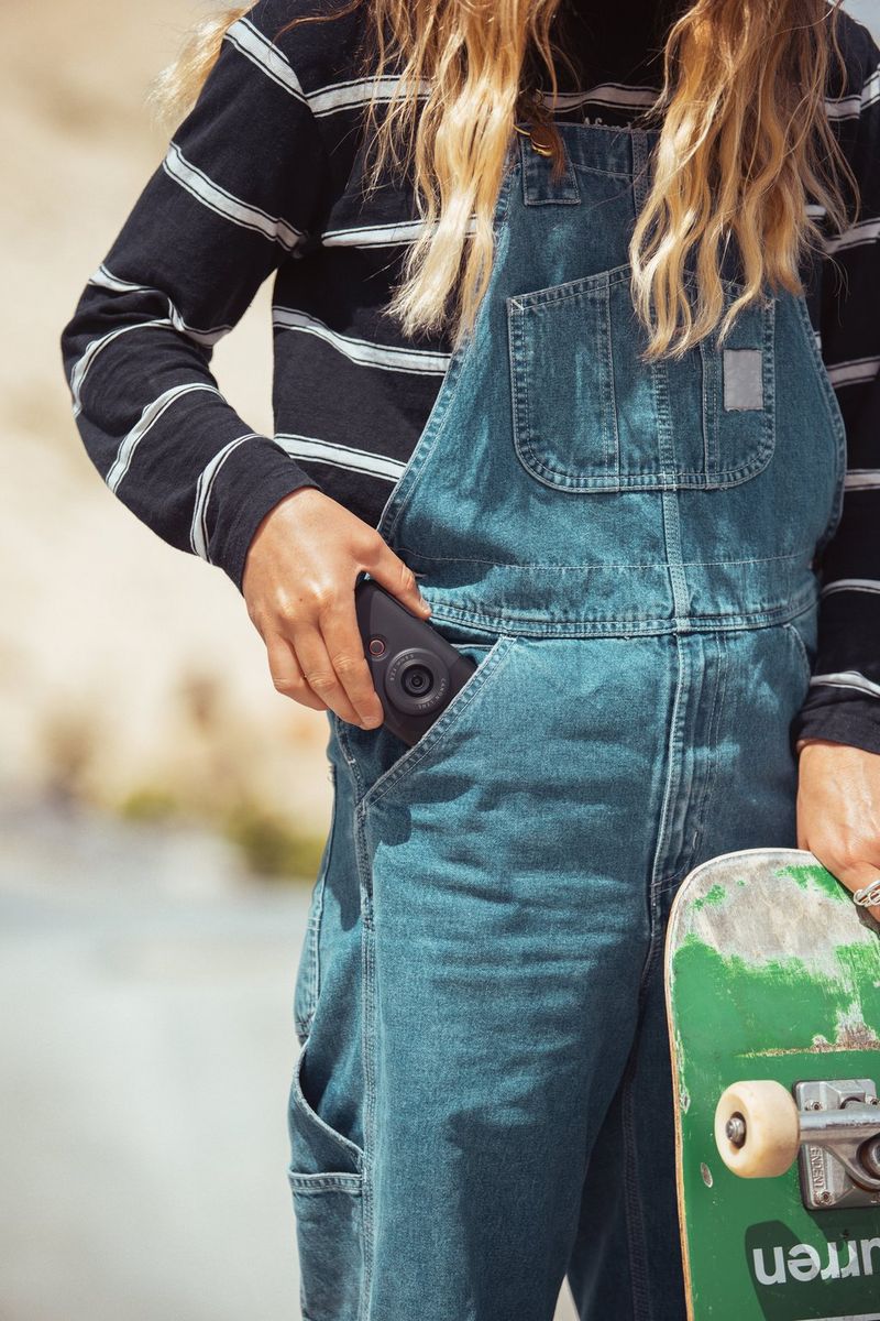 A young woman holding a skateboard puts a Canon PowerShot V10 into the pocket of her dungarees.