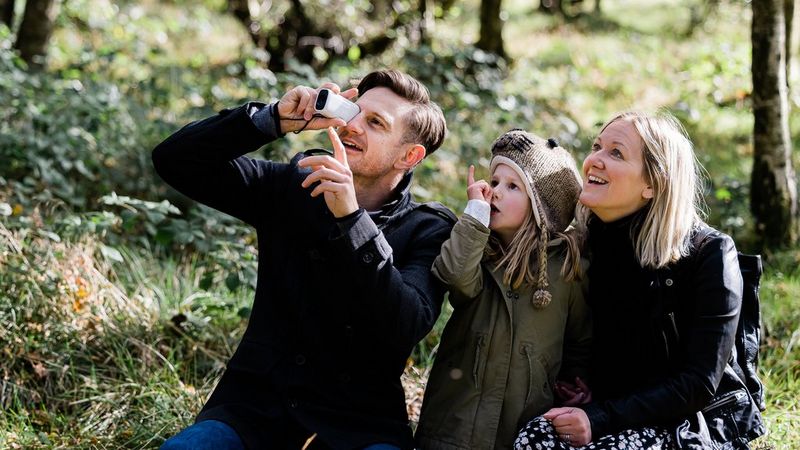 A young girl in a woolly hat sits with her parents in the woods, all three looking at something out of shot as her father uses a Canon PowerShot ZOOM.