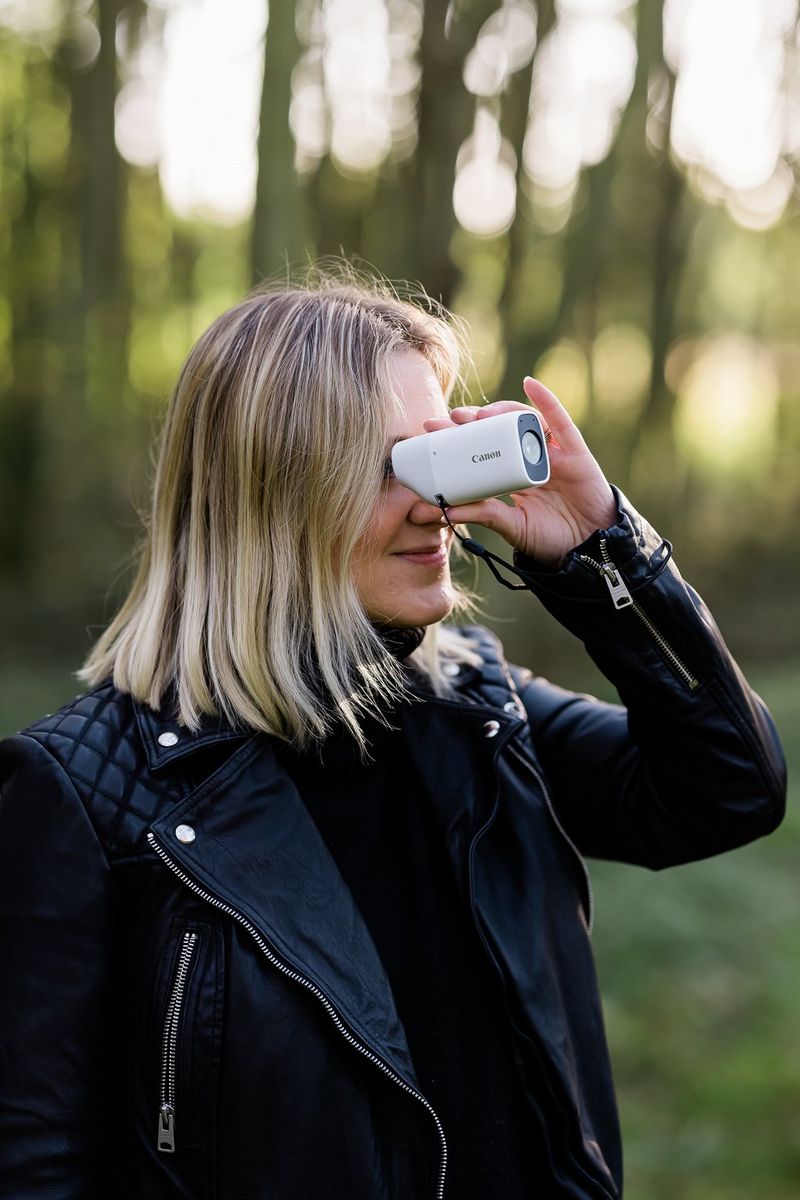 A woman in a leather jacket looks through the viewfinder of a Canon PowerShot ZOOM camera.