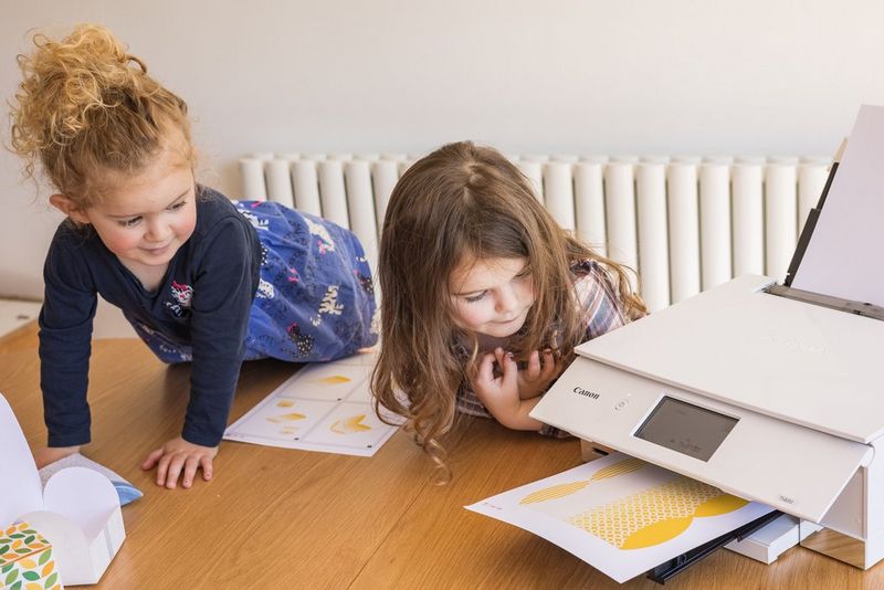 Two young girls eagerly leaning to see a paper box design being printed out of a Canon PIXMA printer.