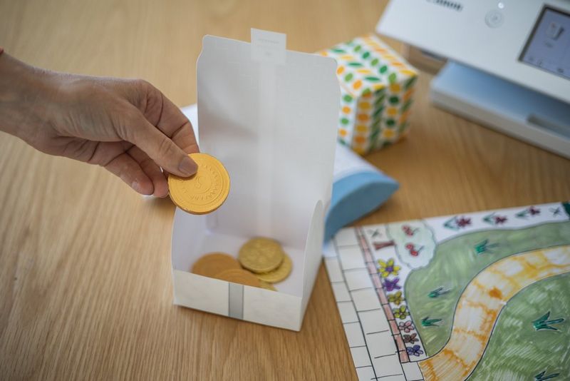 Gold chocolate coins being placed in a papercraft box. Next to the box is a colourful, hand-drawn map.