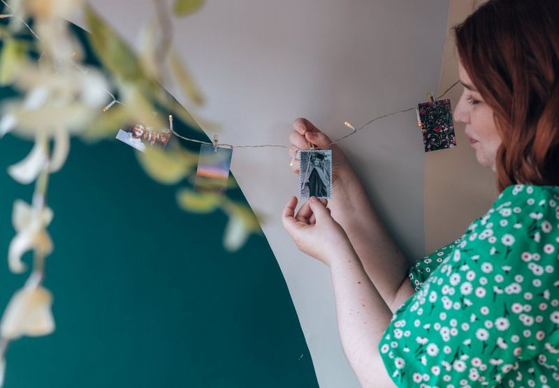 A woman clips a small black and white photo to a string of fairy lights, which has other photographs already clipped along it.