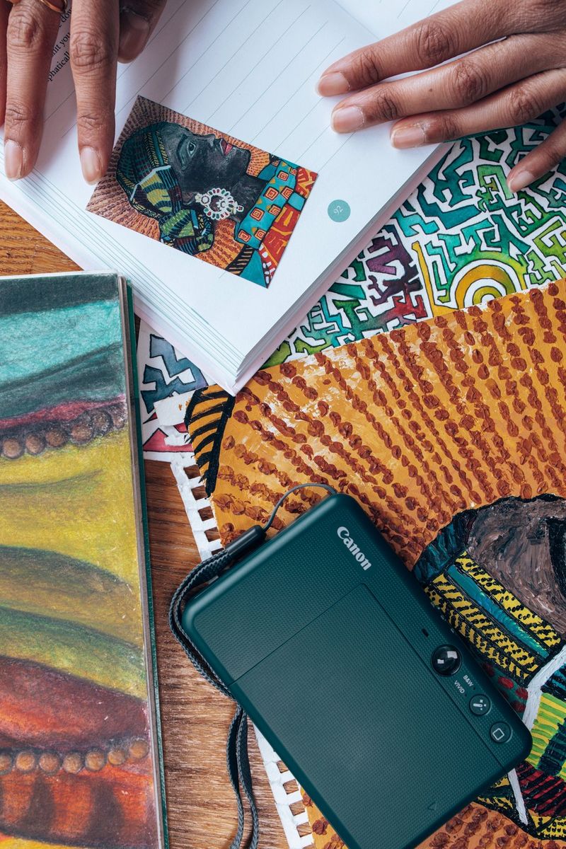 A Canon Zoemini S2 sits face down on pieces of colourful artwork, next to a small sticker of the art being placed into a journal.