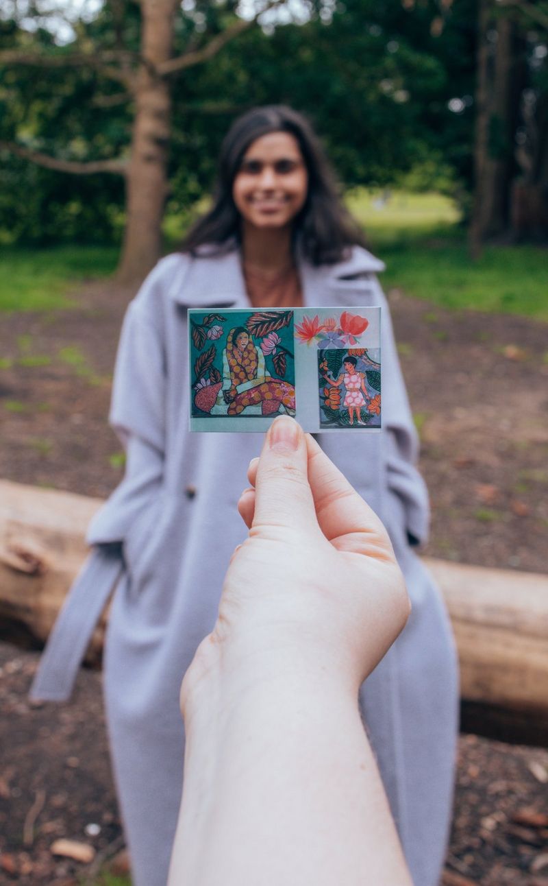 A woman in a grey overcoat smiles at the camera, while a hand holds up a small print of artwork photos in front of her.