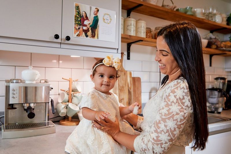 A woman holds an infant on a kitchen surface. A photo calendar is mounted on the cupboard behind her.