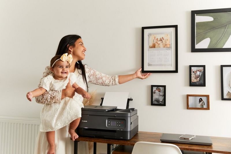 A woman carrying an infant, hanging a framed photo calendar on the wall beside a number of other framed photos and pictures.