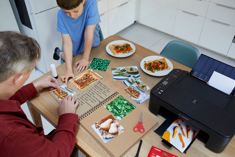 A man and boy sit around a kitchen table which holds a printer and large homemade recipe book, surrounded by printed pictures of food. 