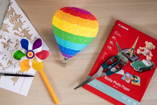 A flat lay image of a wooden desk with a colourful papercrafted windmill and hot air balloon, plus a pack of Canon photo paper.  