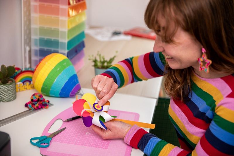 A smiling woman in a colourful striped top sits at a desk finishing off a papercrafted rainbow windmill.