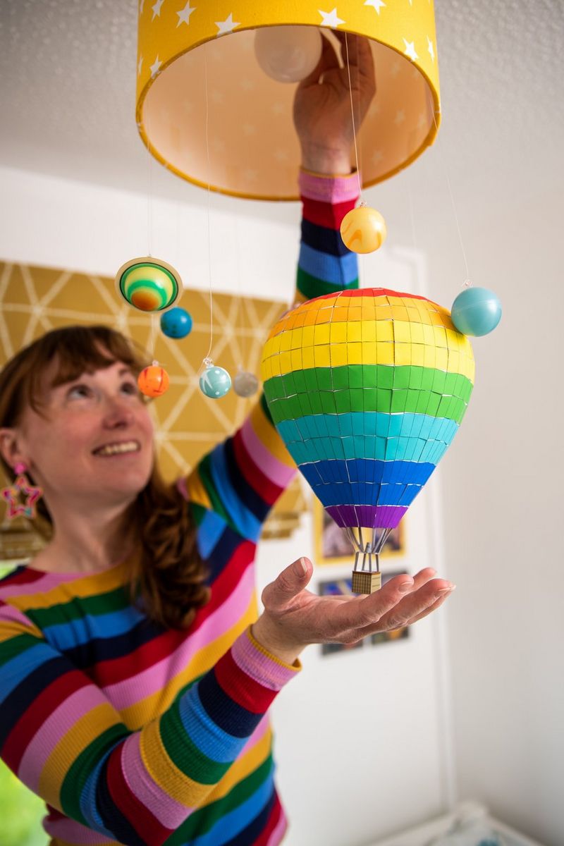 A smiling woman wearing a colourful striped jumper hangs a rainbow-coloured paper hot air balloon from a yellow light fitting. 