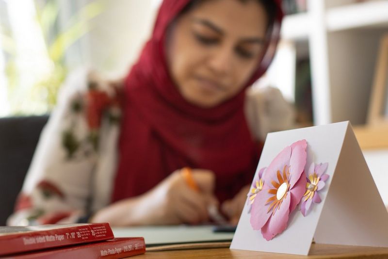 A finished three-dimensional flower card on a table next to two boxes of Canon printer paper. A woman can be seen crafting in the background. 