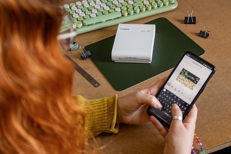 A red-haired woman using the Canon SELPHY Photo Layout app on her phone. A Canon SELPHY Square QX10 printer is on the desk in front of her.
