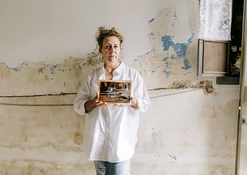 A woman standing in front of a wall holding up a stained black and white photograph of a tricycle.