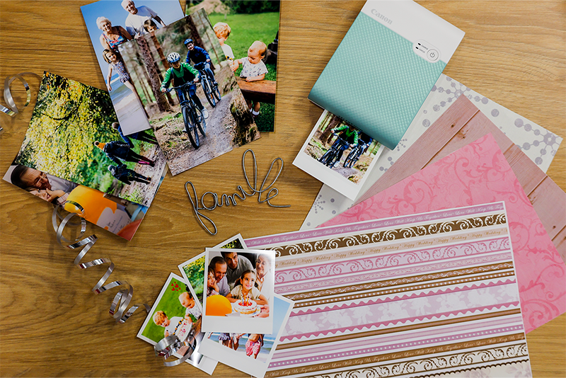 SELPHY Square QX10 printing and image of a family on bikes. On the table there are multiple colourful patterned paper sheets and photos of different sizes of the same family.