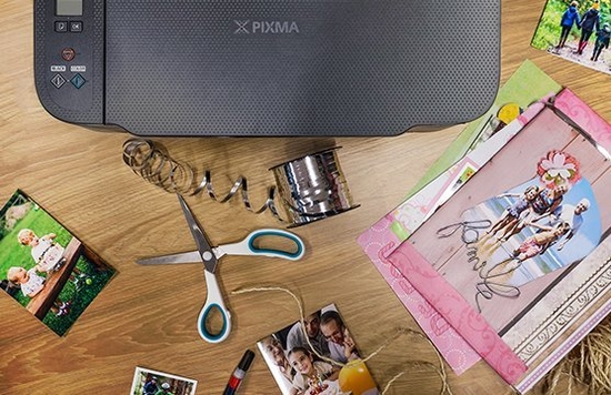 A Canon PIXMA printer on a table with scissors, silver ribbon and pictures surrounding it.