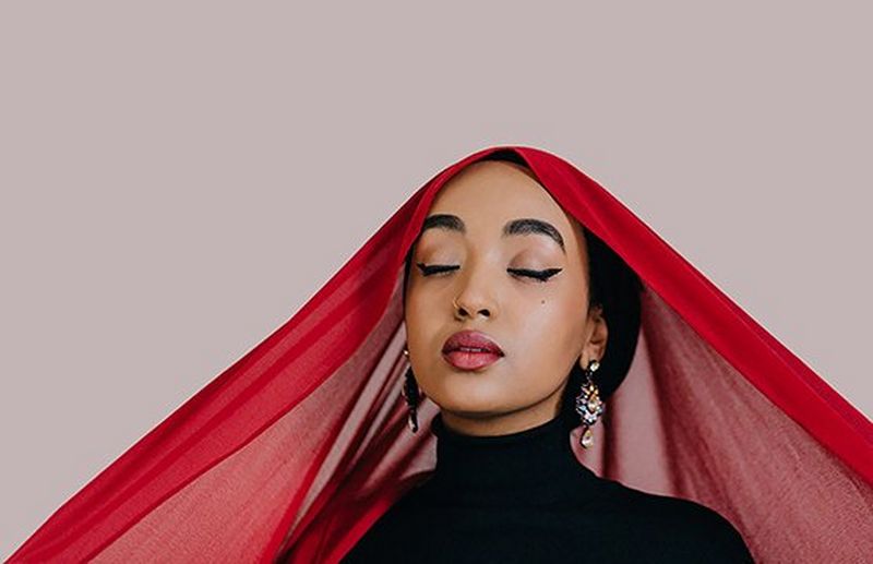 A portrait of photographer and poet Amaal Said with her eyes closed, a flowing red headscarf pulled wide over her shoulders.