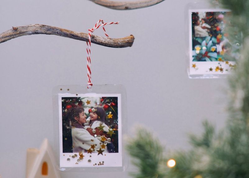 Two Christmas photo decorations, adorned with sequins and printed on a Canon SELPHY Square QX10, hang from branches near a Christmas tree.