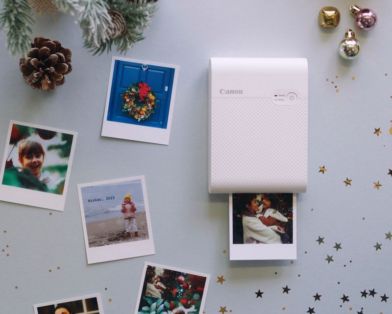 A flatlay showing a selection of Christmassy photo prints, a Canon SELPHY Square QX10 printer and festive decorations.