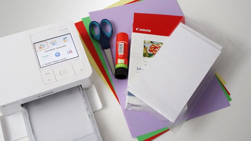 A Canon SELPHY printer sits next to a pile of colourful cardboard, a pack of Canon printer paper, a pair of scissors and a glue stick.