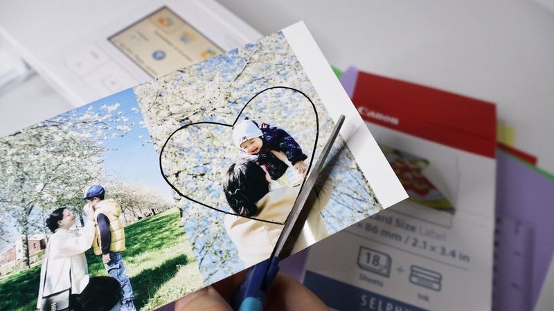 Scissors being used to cut out a heart shape around a printed photograph of a woman holding up a small child. In the background is a Canon SELPHY printer and a pack of Canon photo paper.