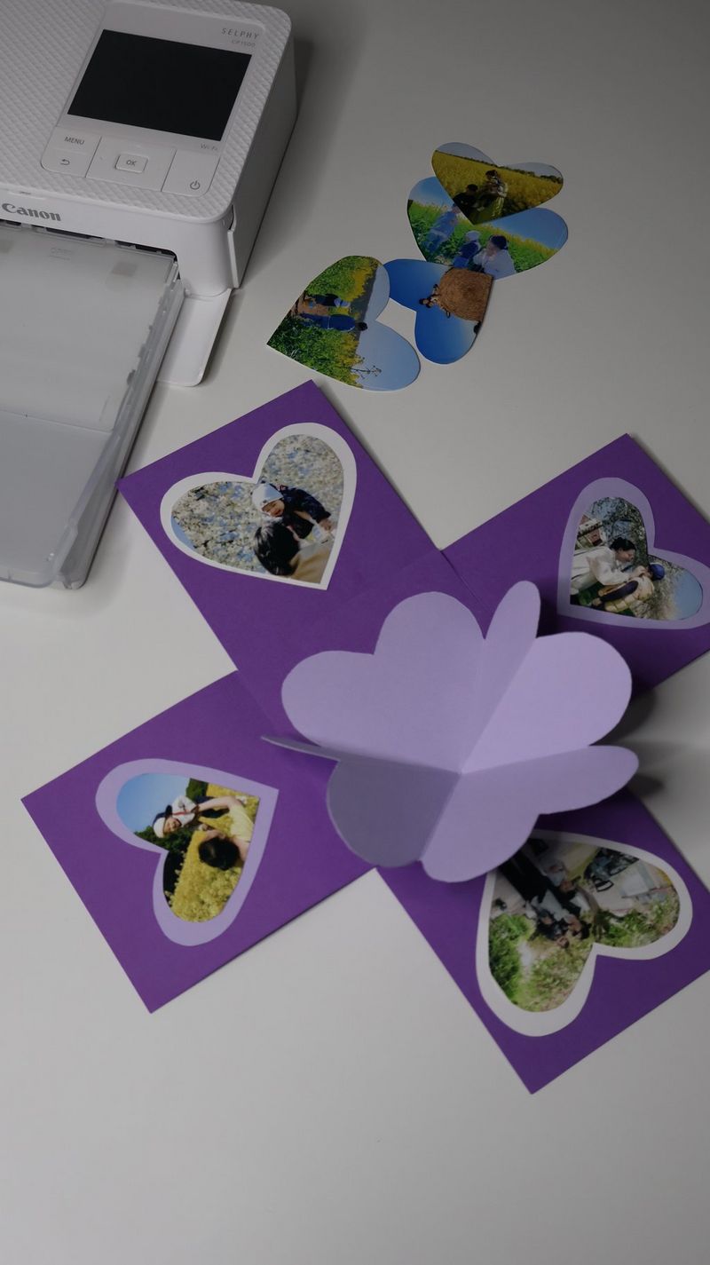 A cross-shaped piece of purple cardboard sits on a worktop, with heart-shaped photographs stuck onto it and a pop-up heart in the middle. A Canon SELPHY printer sits near it.