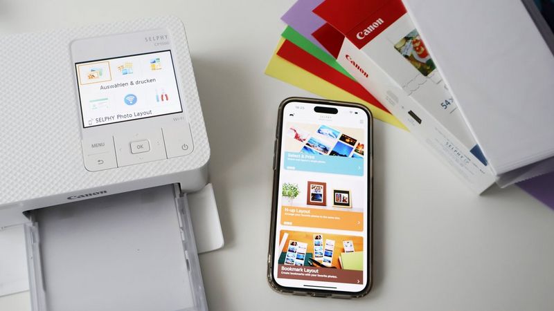 A Canon SELPHY printer sits next to a smartphone showing the Canon SELPHY Photo Layout app, some coloured cardboard and a pack of Canon photo paper.
