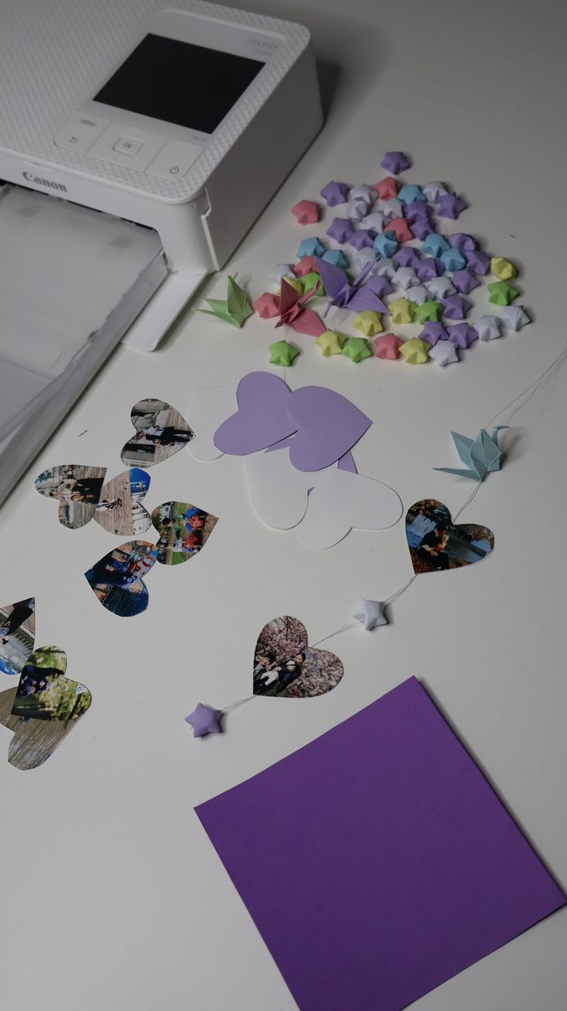 A piece of purple cardboard sits on a worktop, with heart-shaped photographs and papercraft stars next to it. A Canon SELPHY printer sits near it.