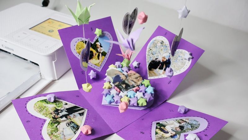 A purple cardboard explosion box filled with papercraft stars, decorated with heart-shaped photographs and has more photographs and origami hanging from strings above it. A Canon SELPHY printer sits near it.