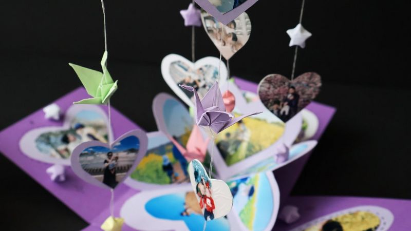A purple cardboard explosion box filled with papercraft stars, decorated with heart-shaped photographs and has more photographs and origami hanging from strings attached to the lid held above it.