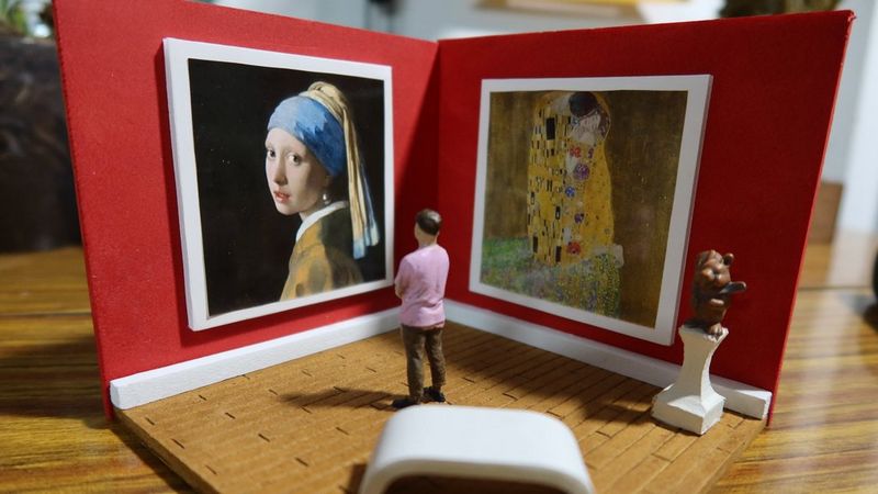A mini papercraft art gallery with two paintings hung on red walls, and a mini figure standing in the foreground, looking at the art.