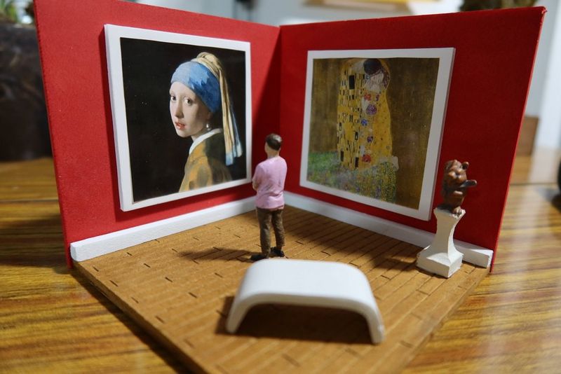 Umelecká galéria mini výtvorov z papiera s dvoma maľbami zavesenými na červených stenách a mini postavou v popredí, ktorá sa pozerá na dané umenie.