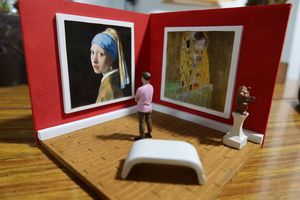 Miniatūra papīra mākslas galerija ar divām gleznām, kas piekārtas pie sarkanām sienām, un miniatūru figūru, kas stāv priekšplānā un skatās uz mākslas darbiem.
