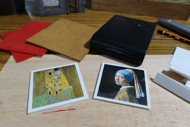 Prints of Girl with a Pearl Earring by Johannes Vermeer and The Kiss by Gustav Klimt lay on a table in front of a Canon SELPHY Square QX10 printer and other art materials.