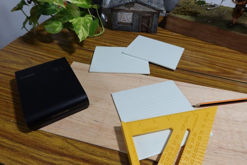 A Canon SELPHY Square QX10 is positioned on a tabletop next to pieces of hard foam, a ruler and a pencil.