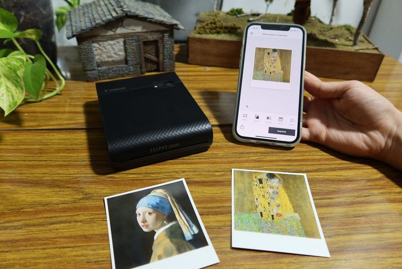 A hand holds a smartphone with the Canon SELPHY Photo Layout app on the screen, displaying The Kiss by Gustav Klimt, with a printout of the painting and Girl with a Pearl Earring by Johannes Vermeer, as well as a Canon SELPHY Square QX10, on the table below.