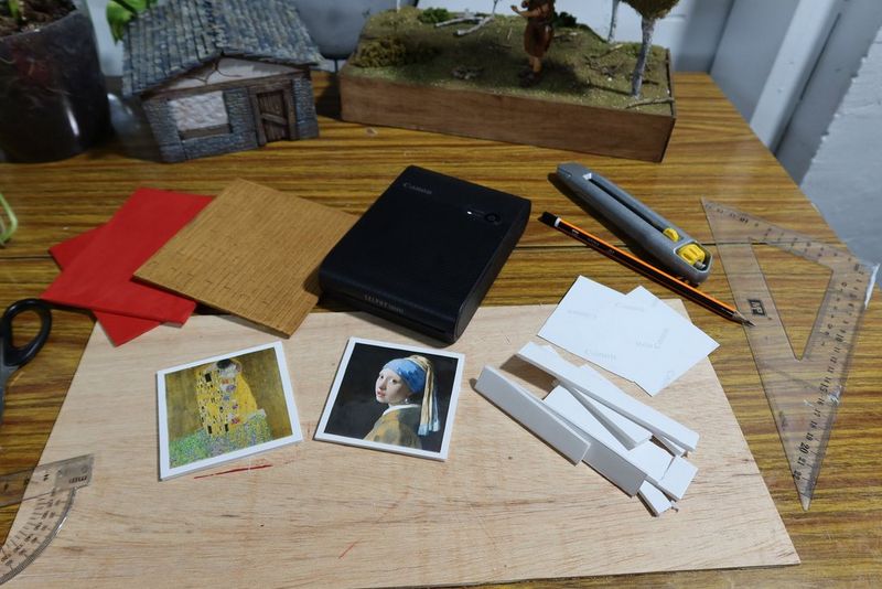 Materials for the projects are laid out on a desktop, including painted hard foam, a Canon SELPHY Square QX10 printer, two mini prints of paintings, a pencil, ruler, penknife and excess foam.
